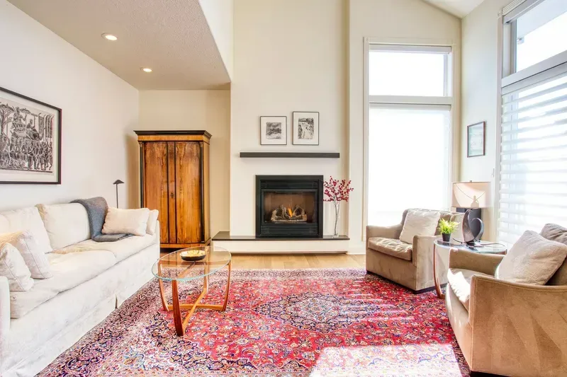 Living room with fireplace, white walls, wood floors, and red rug; sofa, chairs, and wooden cabinet.