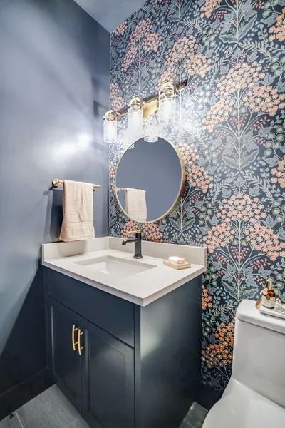Powder room with floral wallpaper, navy blue vanity, gold mirror and lighting.