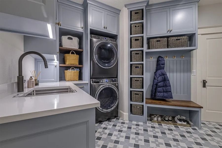 Laundry room with blue cabinets, stacked washer/dryer, sink, baskets, and bench.