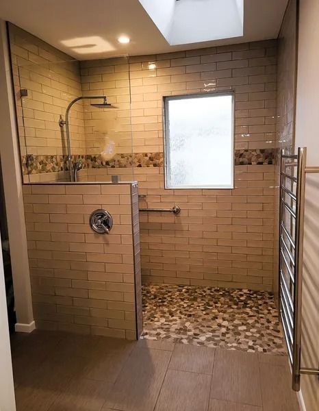A tiled walk-in shower with pebble floor, glass enclosure, and a window. Includes a towel warmer and hand bar.