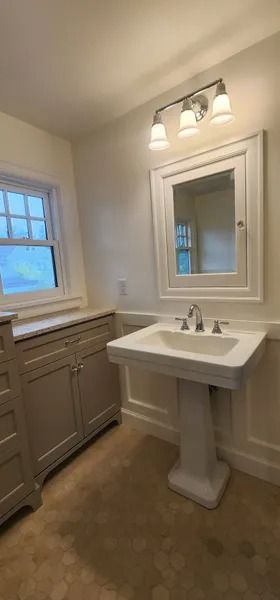 Bathroom with pedestal sink, cabinet, and window. White and neutral colors, tile floor.
