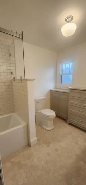 Bathroom with a bathtub, toilet, and two cabinets in a light-toned color scheme with a window.