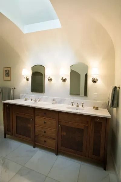 Bathroom with wooden vanity, marble countertop, two arched mirrors, sconces, and skylight.