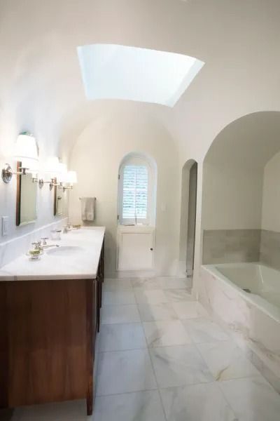 White bathroom with vaulted ceiling, marble floors, wood vanity, and a skylight.