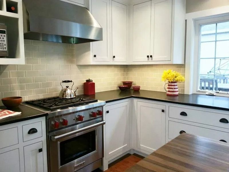 White kitchen with stainless steel oven, backsplash, and cabinets, black countertops, and wooden table.