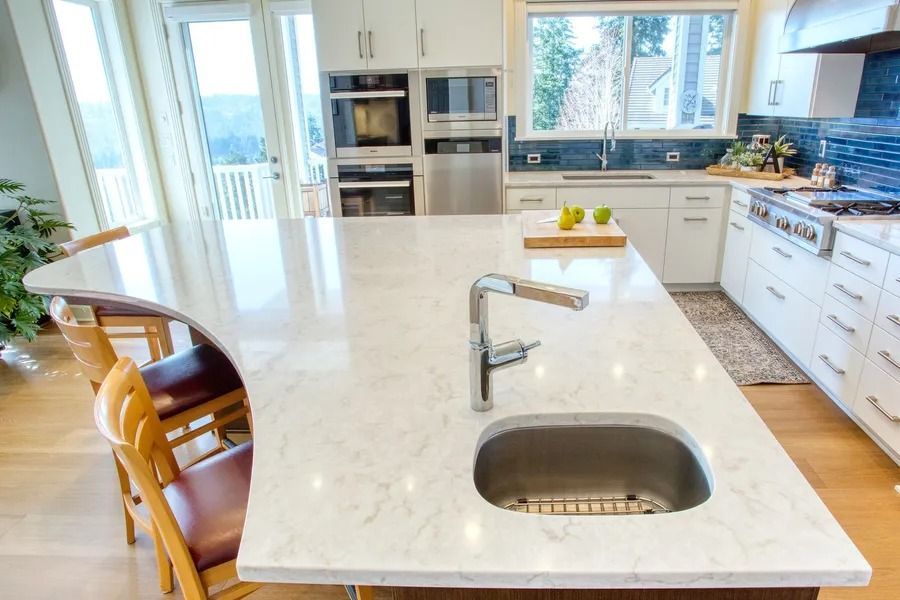 Modern kitchen with white countertops, island with sink, stainless steel appliances, and wood floors.