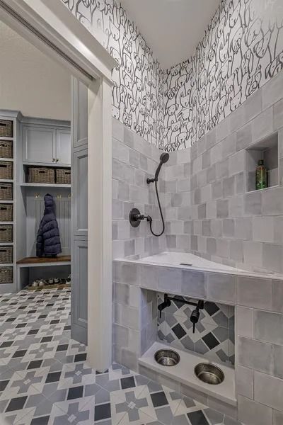 Dog shower with gray tile, showerhead, dog bowls, and patterned wallpaper. A mudroom is visible in the background.