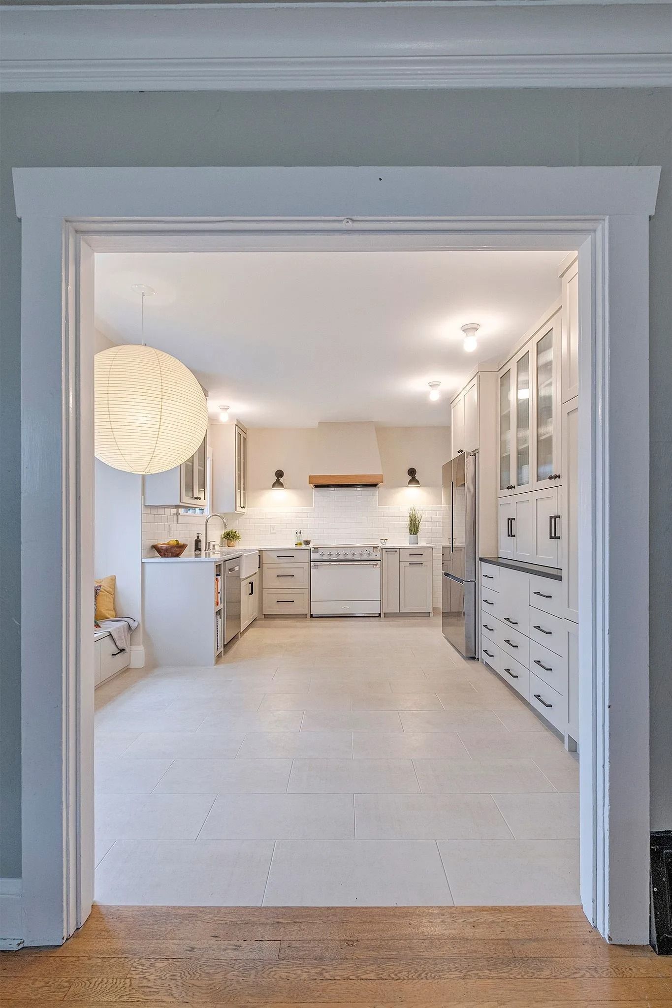 View of a bright, white kitchen from a doorway. White cabinets, appliances, and a hanging light fixture are visible.
