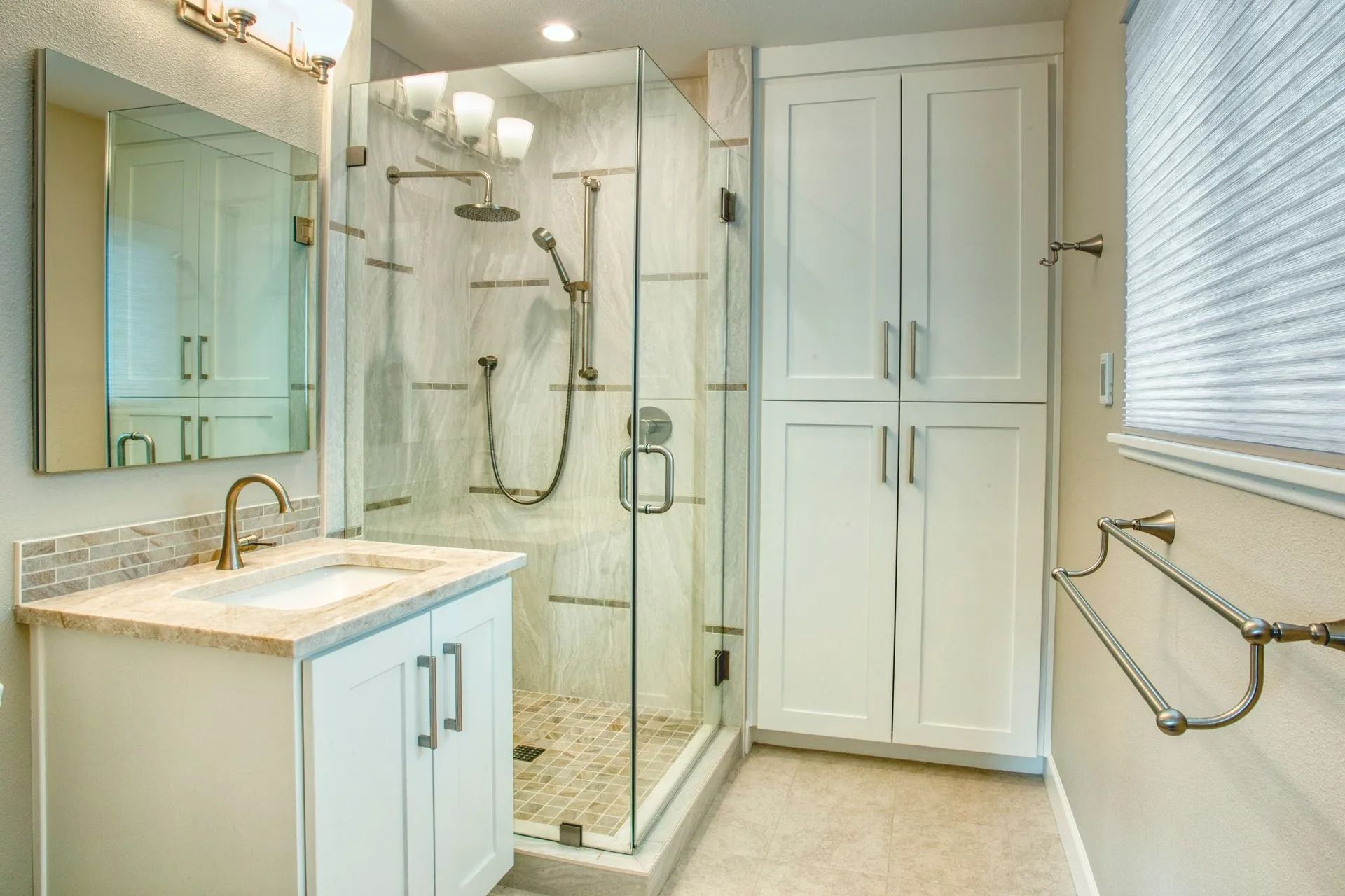 Bathroom with white cabinets, glass shower, and beige countertop, next to a tall storage cabinet.