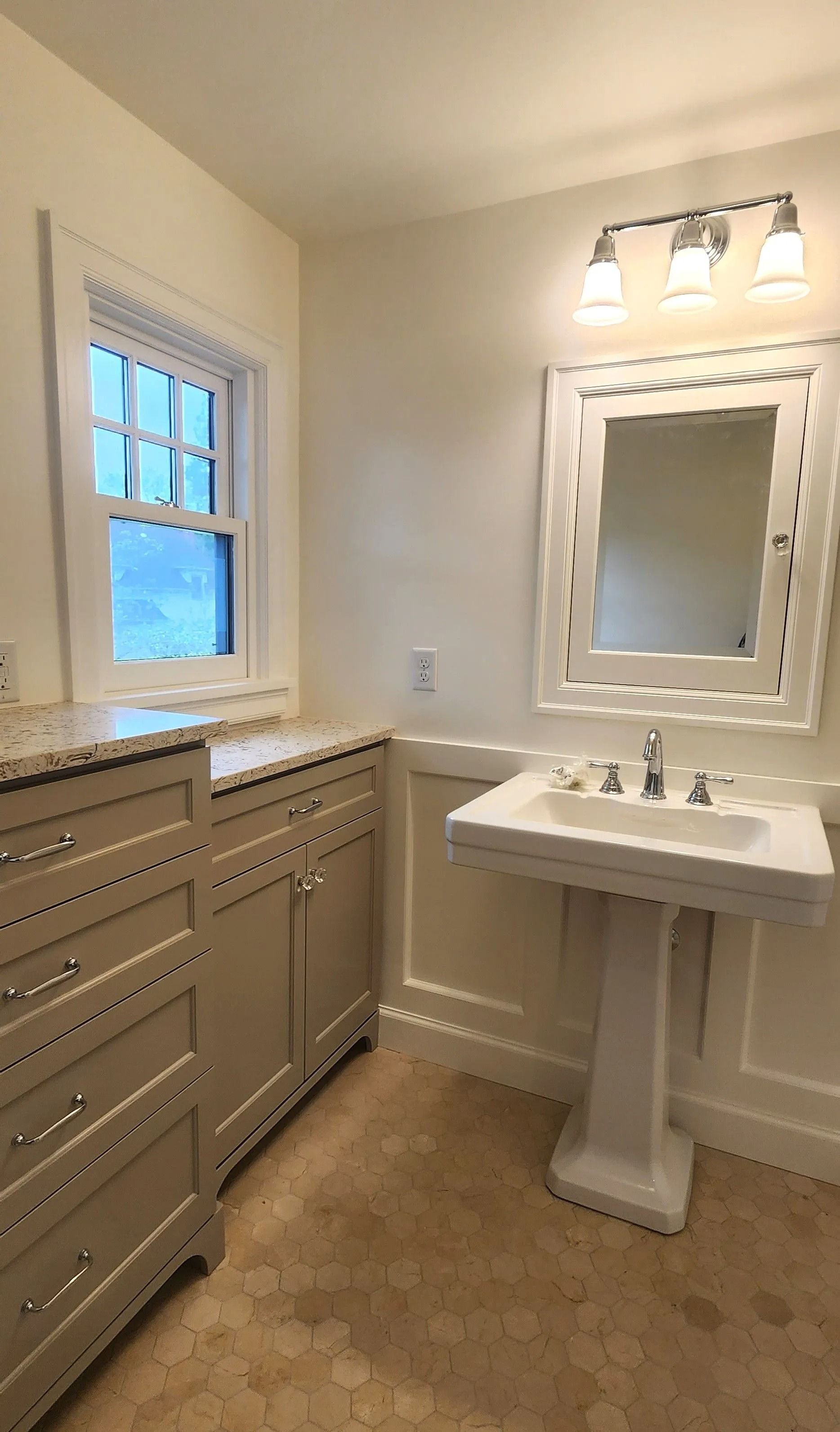 Bathroom with pedestal sink, vanity, window, and mirror with light fixture. Beige walls, beige floor.