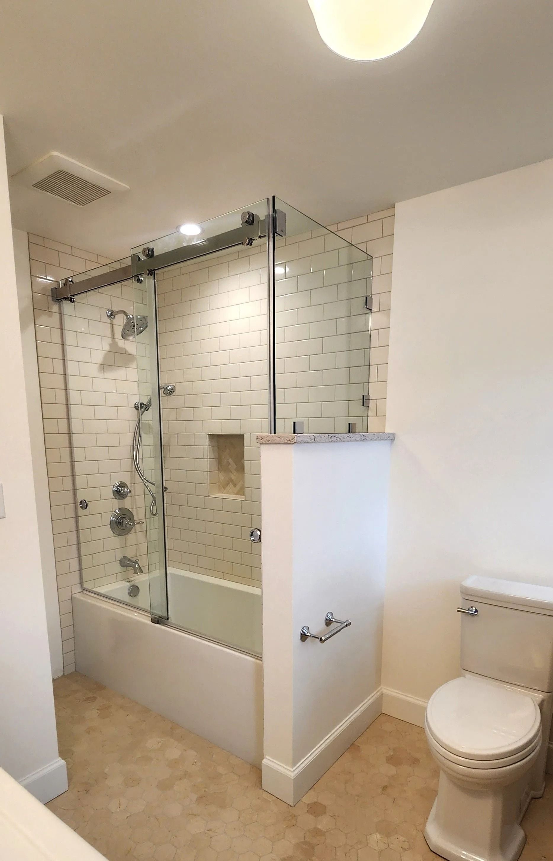 Bathroom with tub and shower behind a glass enclosure. White tile, tan flooring, and a toilet.