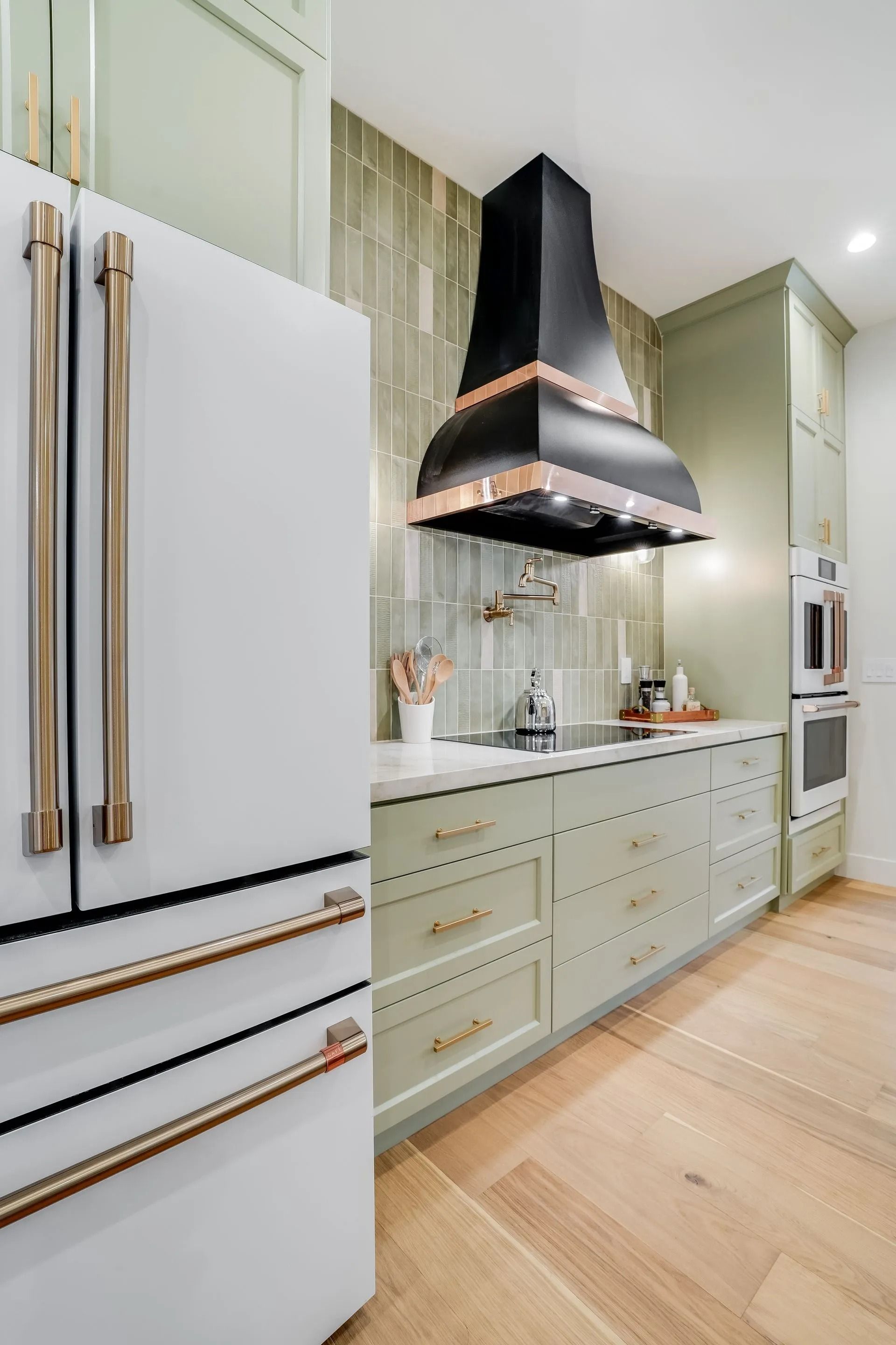 Green kitchen with white refrigerator, black range hood, and light wood flooring.