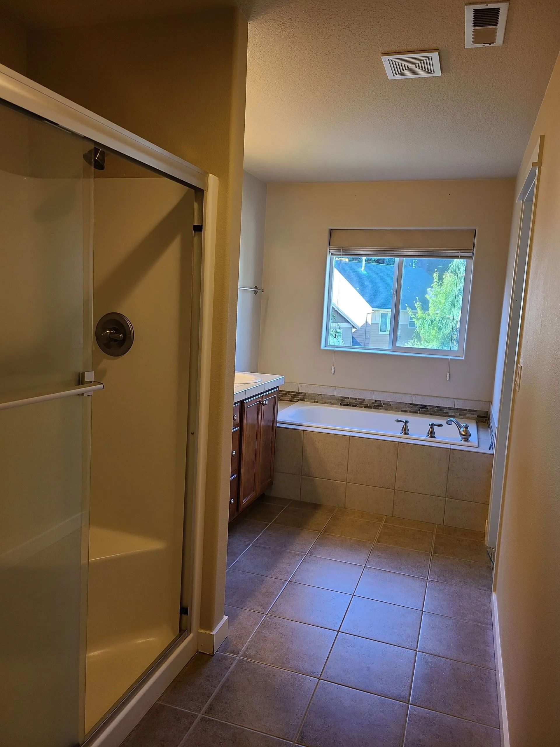 Bathroom with shower on left, vanity and window with bathtub in background. Beige walls, brown floor.