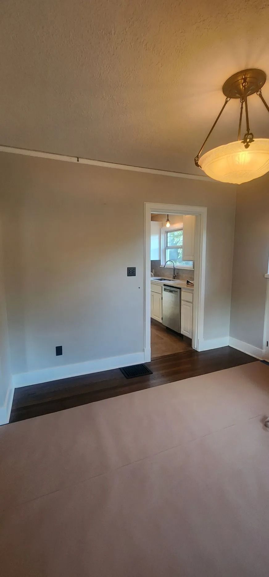 Interior of a room with tan walls, white trim, and a doorway to a kitchen. Brown hardwood floor. Beige carpet.
