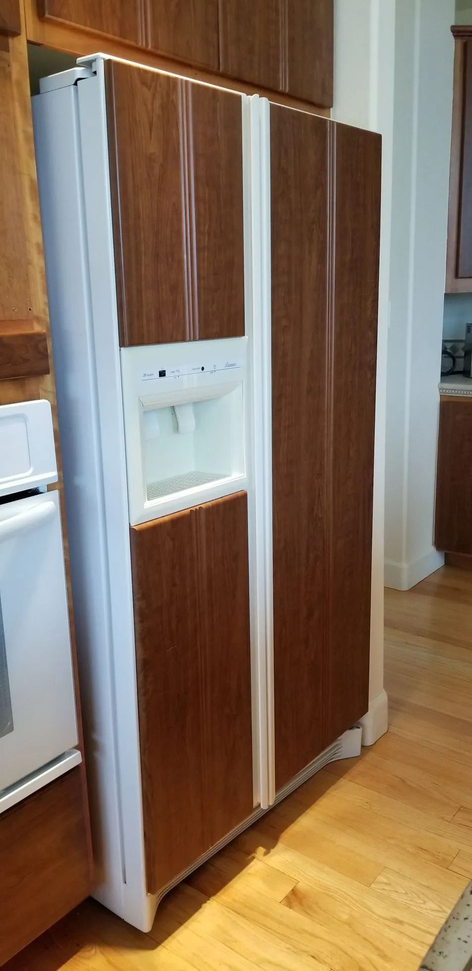 Side-by-side refrigerator with white trim and wood-look doors. Built-in ice and water dispenser. Kitchen setting.