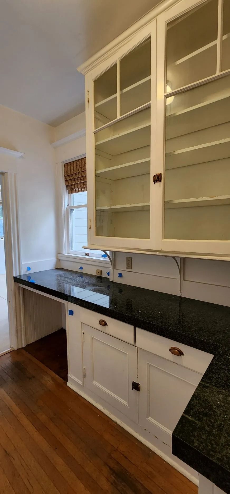 Kitchen with white cabinets, dark countertop, and wooden floor. Window with roman shade.