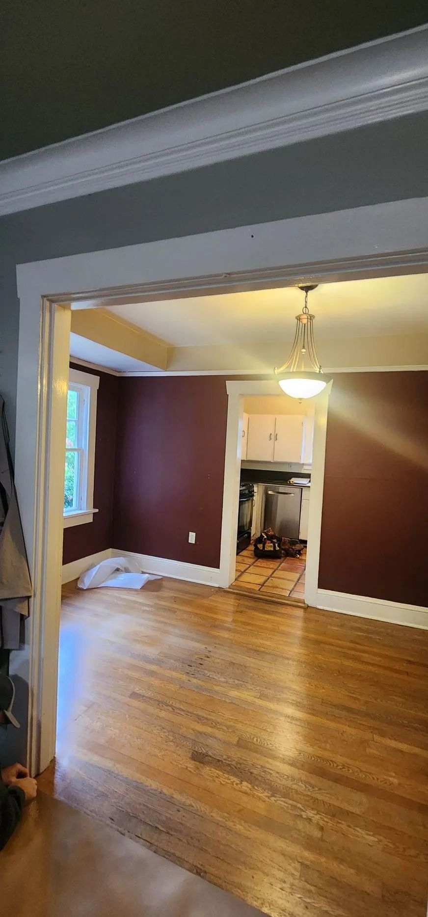 Interior view of a house. Hardwood floors, maroon walls, and a view into the kitchen. White trim, and a light fixture.