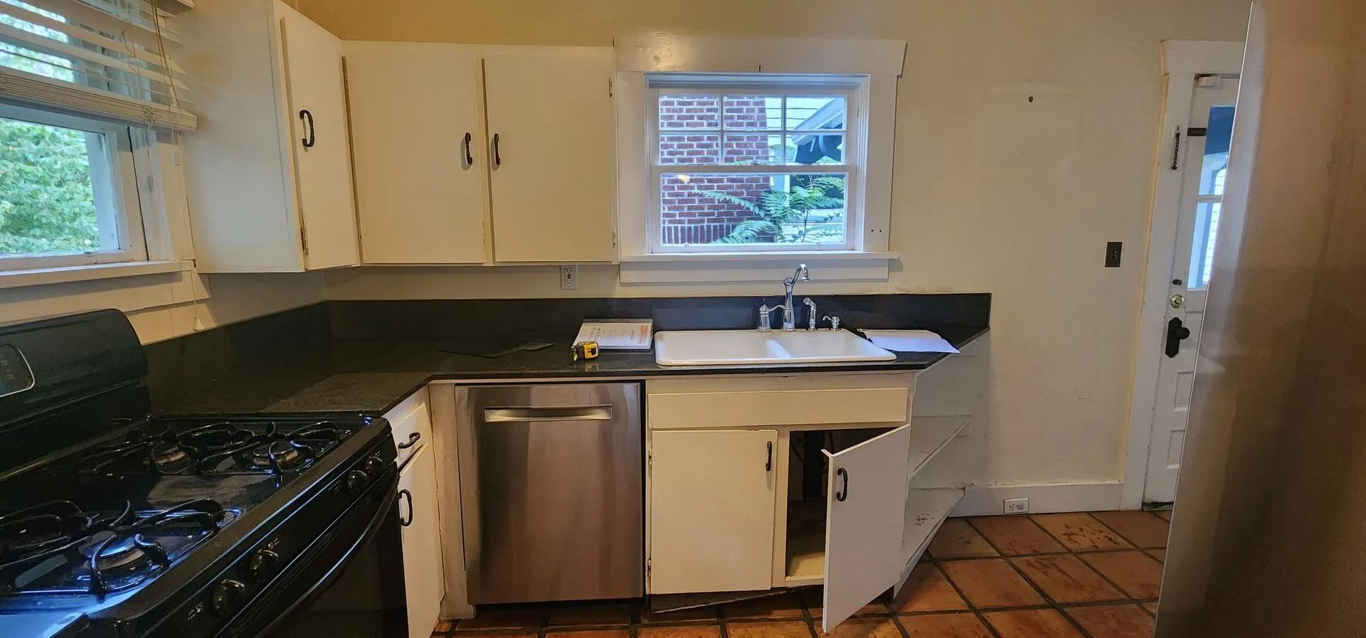 Kitchen with white cabinets, dark countertops, a stainless steel dishwasher, and a window with a view of greenery.