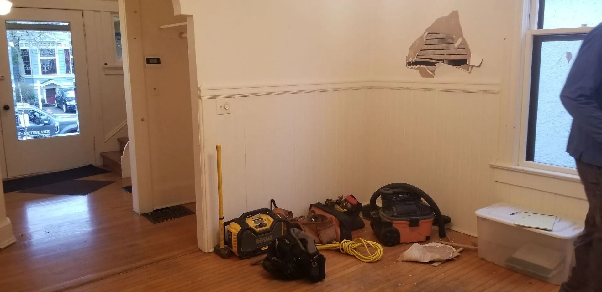 Room interior with damaged wall, tools, and a person standing near a window. A door with glass is visible.