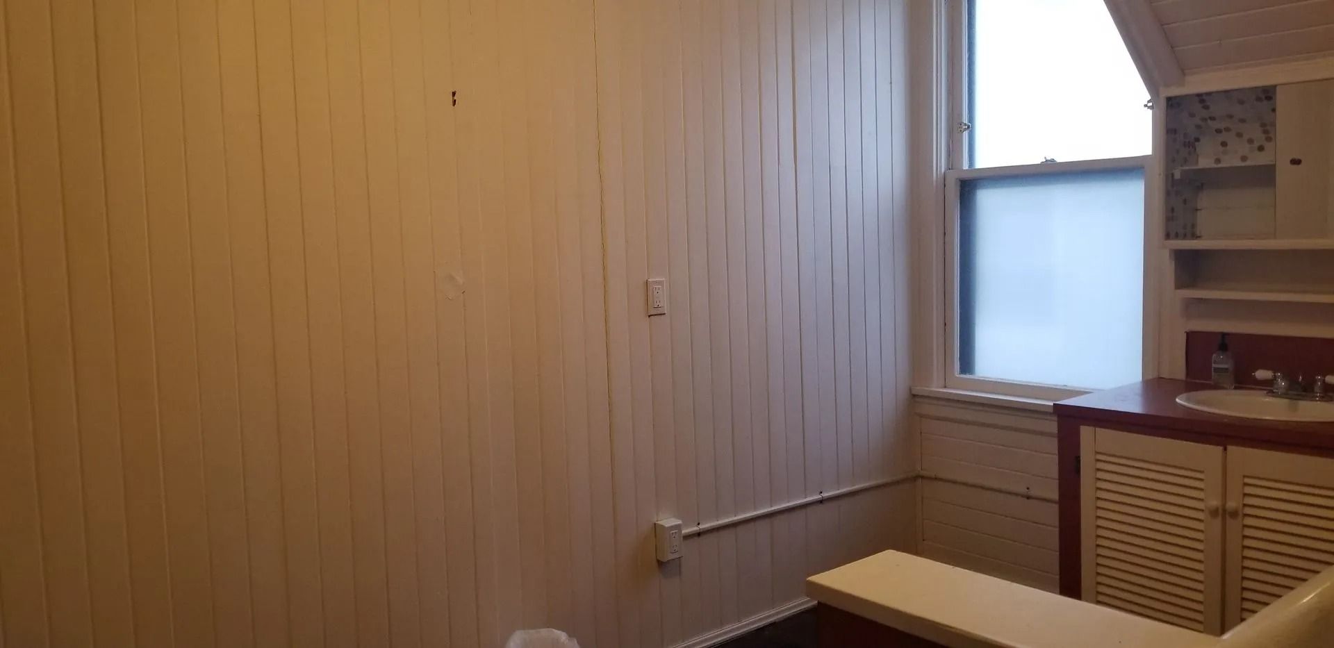 A bathroom with wood-paneled walls and a window. A white countertop with a sink is visible near the window.