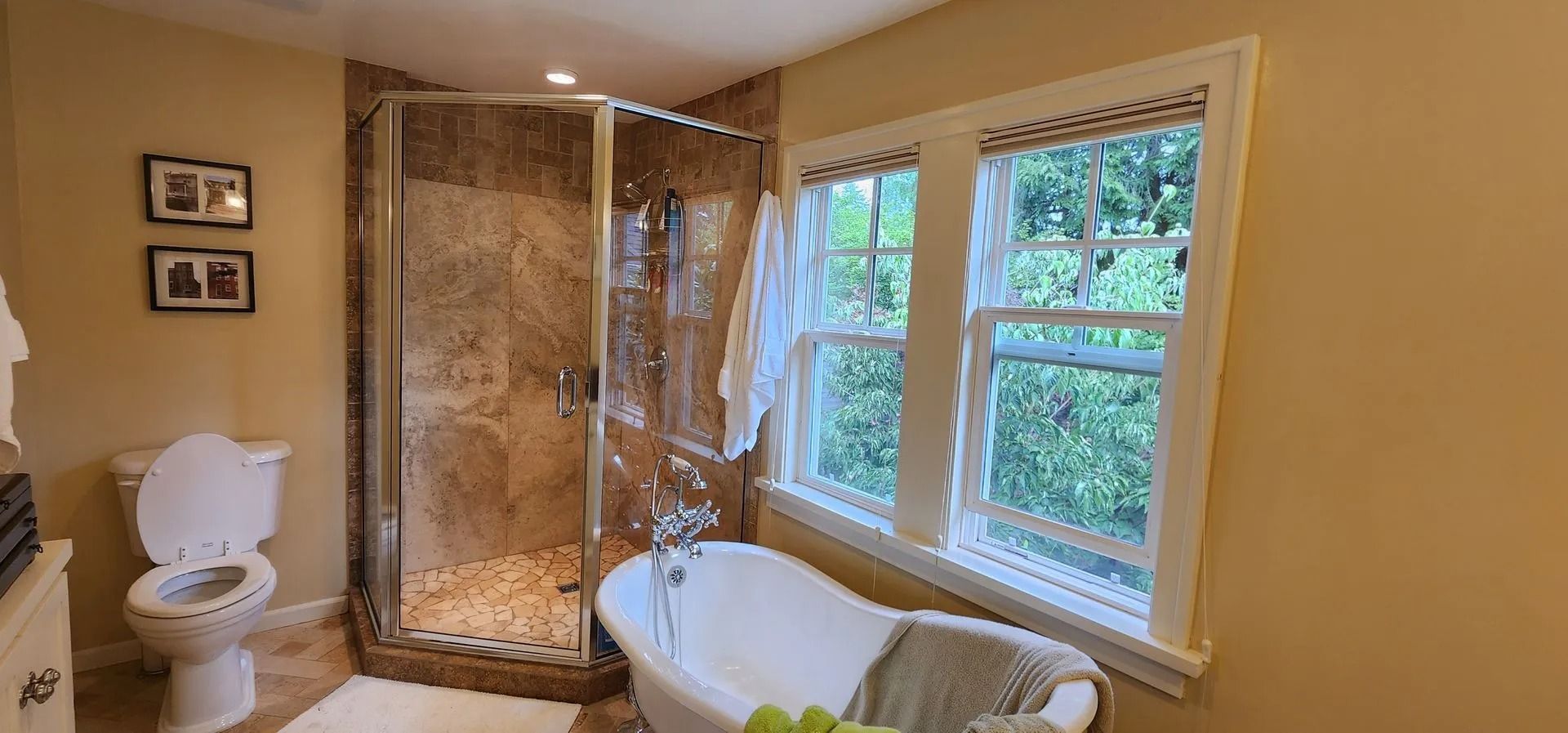 Bathroom with shower, tub, toilet, and window with greenery. Warm color palette.