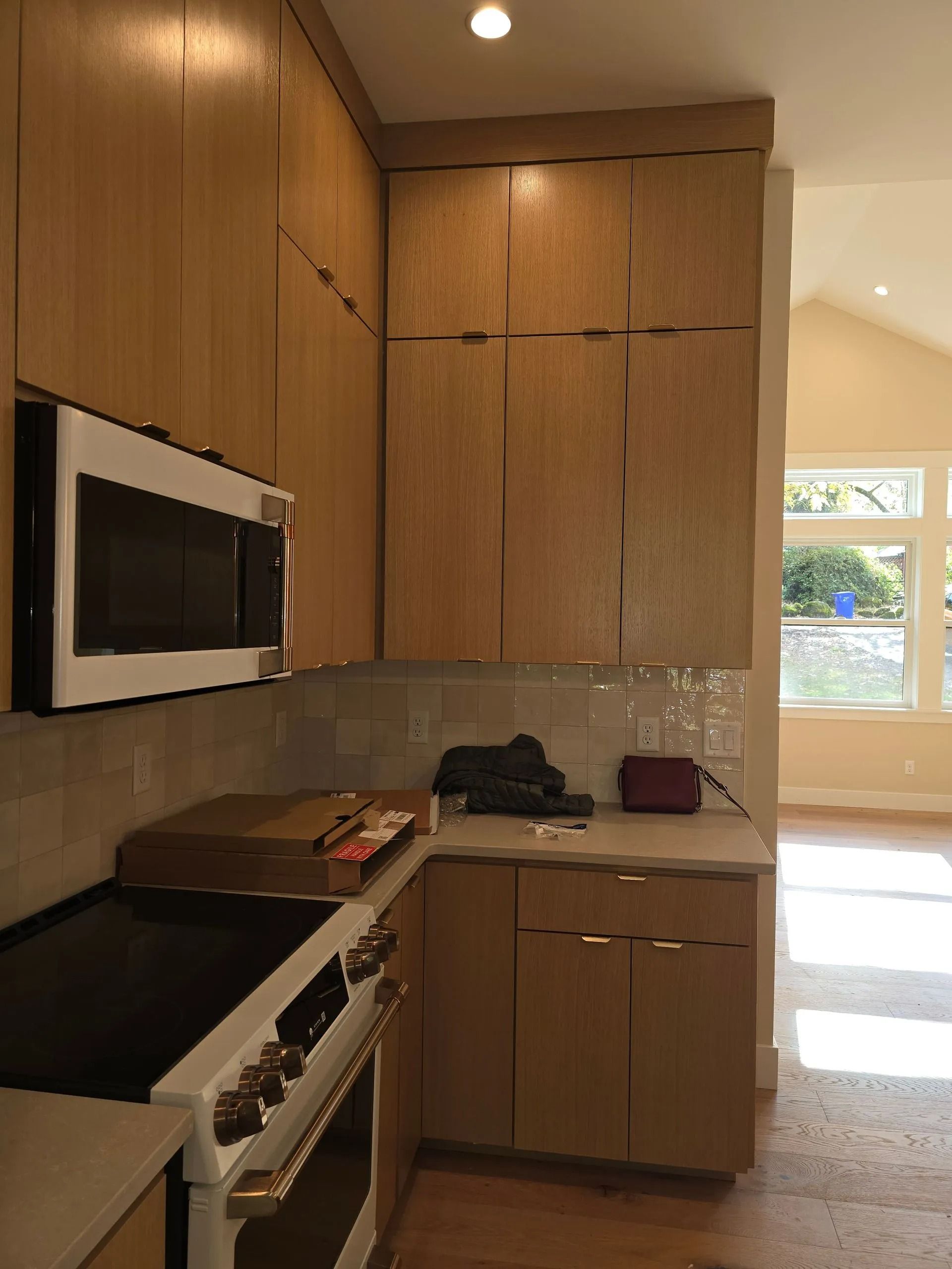 Kitchen corner with light wood cabinets, microwave, stovetop, and a glimpse of a sunlit room.