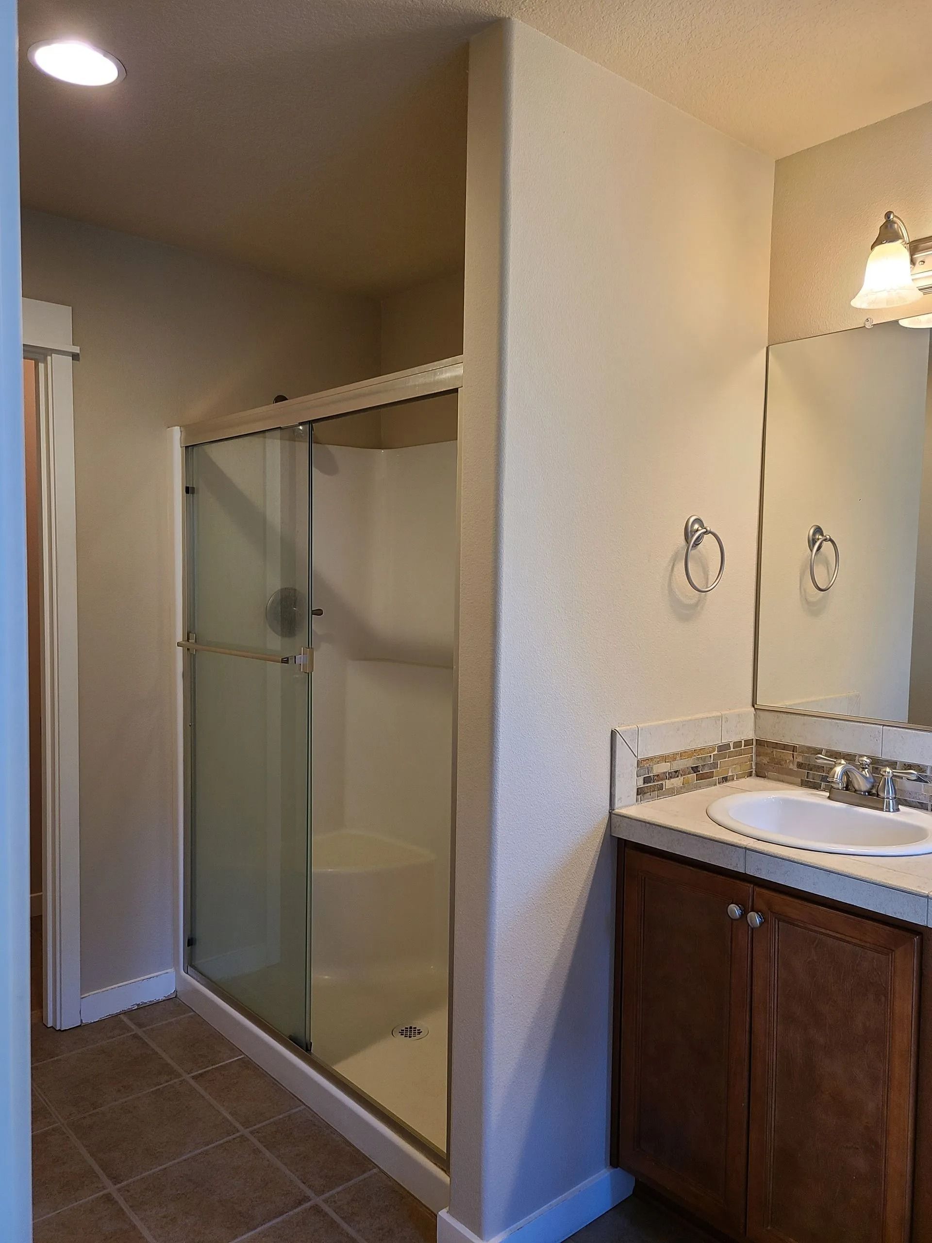 Bathroom with a shower, vanity, and mirror. White walls and brown tiled floor.