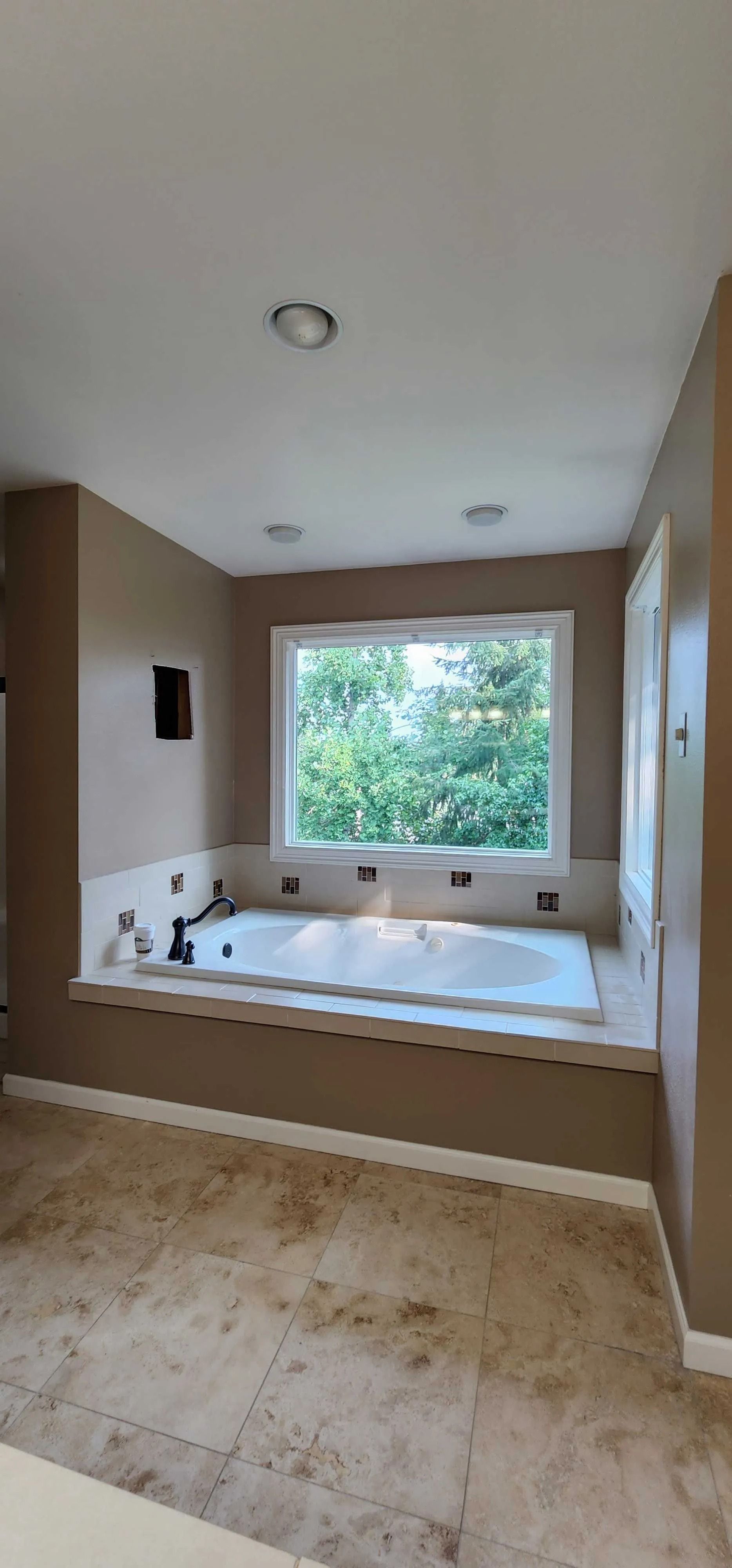 Bathroom with a built-in tub under a window. Beige walls, tile floor, and recessed ceiling lights.