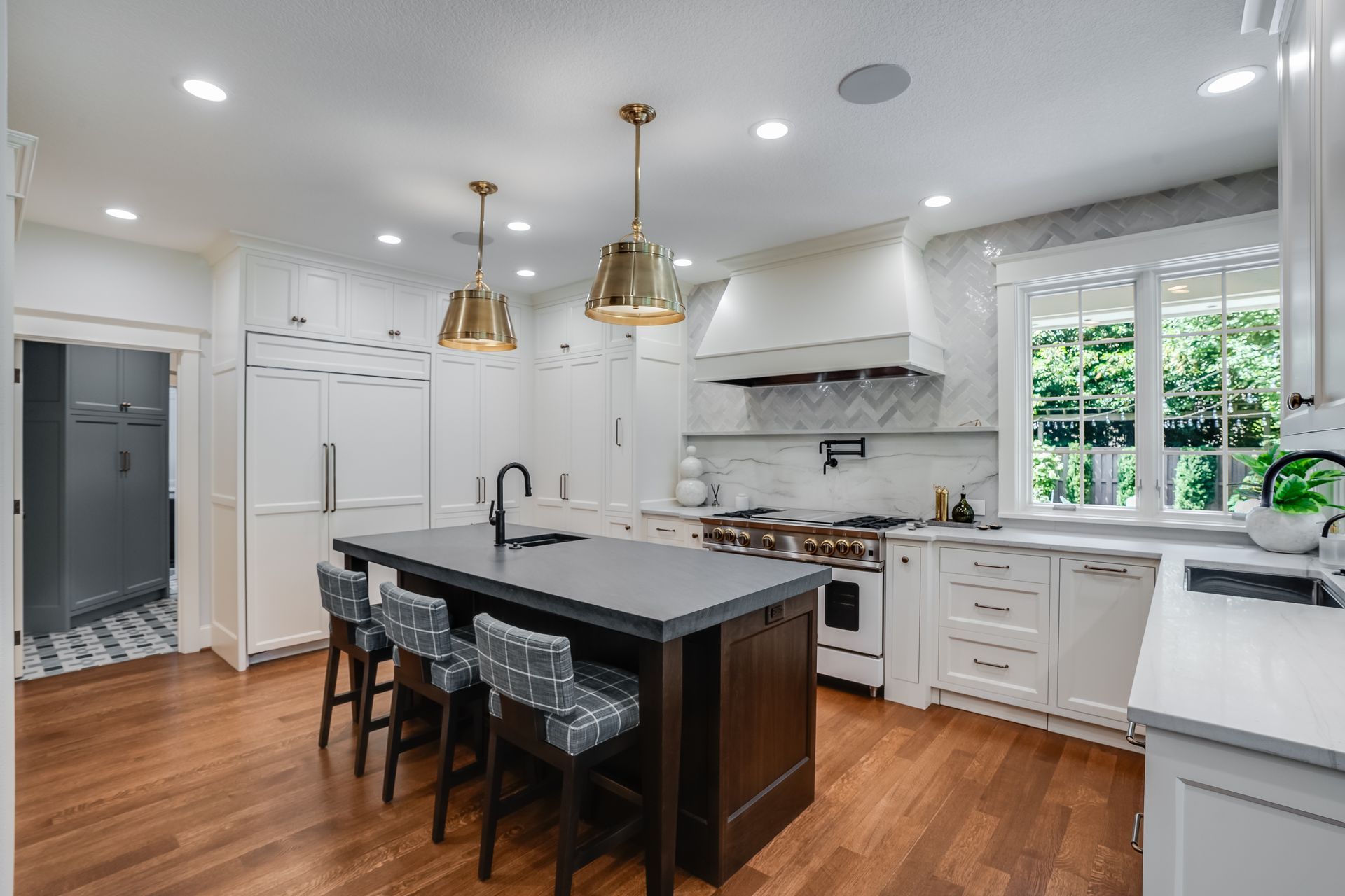 A modern white kitchen featuring a dark wooden island with three stools, gold pendant lights, and hardwood floors.