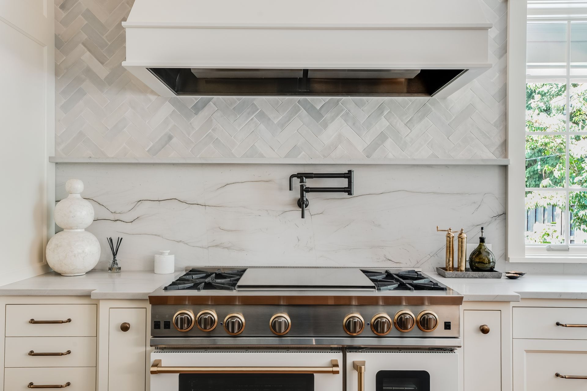 A kitchen range with a marble slab backsplash, herringbone tile above, and a white vent hood in a bright, modern space.