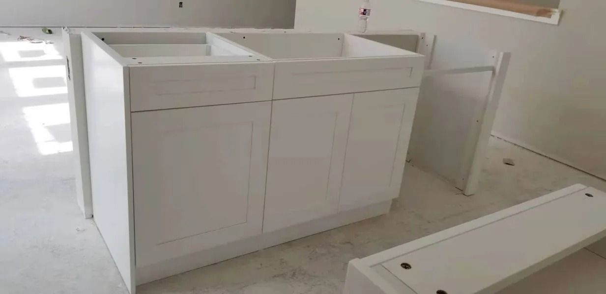 White kitchen cabinets are installed in a room. Some cabinet parts are lying nearby.