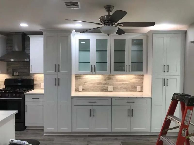 Modern white kitchen cabinets with glass-front uppers and a gray backsplash. A ceiling fan and ladder are present.