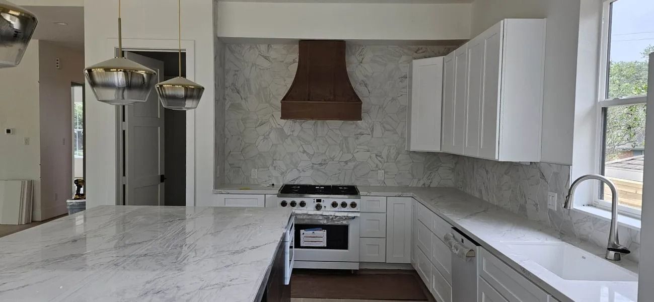 A modern kitchen with a white marble island, cabinets, and backsplash. A wooden hood and range complete the space.