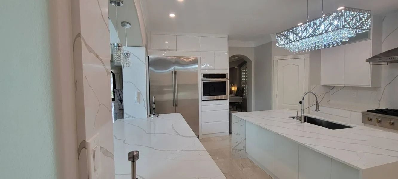Modern kitchen with white countertops, cabinets, and a stainless steel refrigerator. A crystal chandelier hangs above an island.