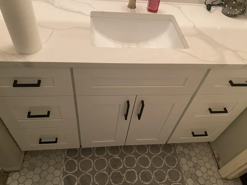 White bathroom vanity with black hardware, a rectangular sink, and a gray patterned rug.