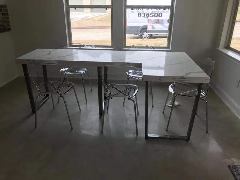 Modern dining table with marble top and metal legs, four clear chairs. Windows in the background.