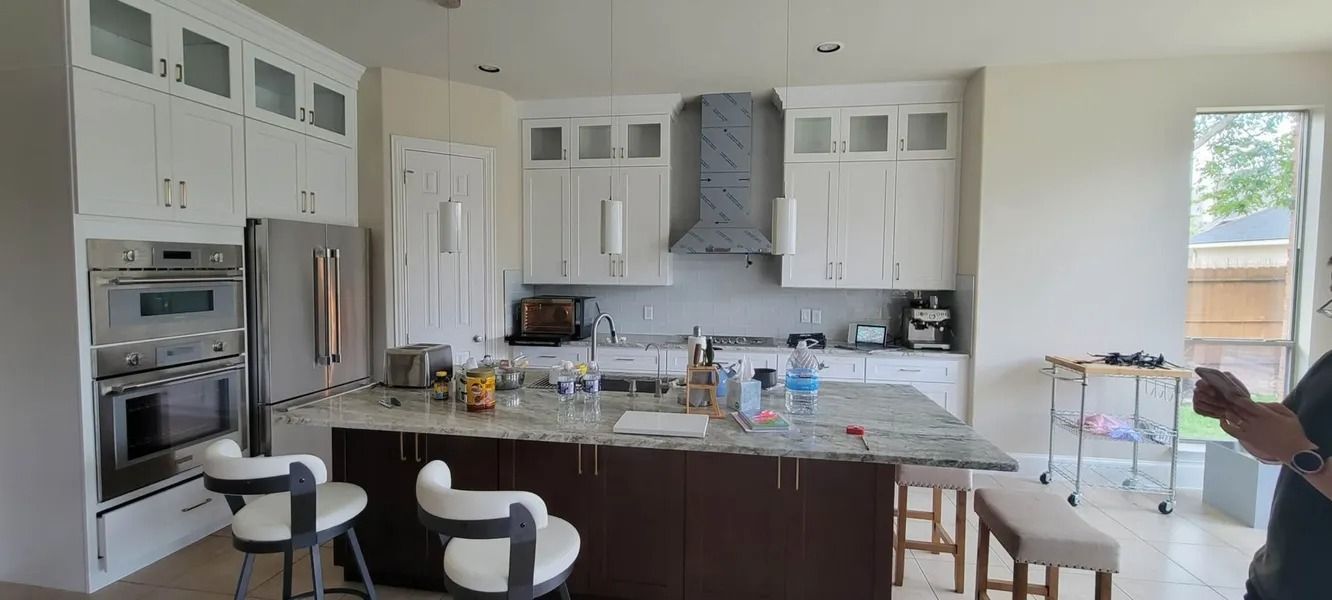 A bright white kitchen with a large island, stainless steel appliances, and a person's arm in the corner.