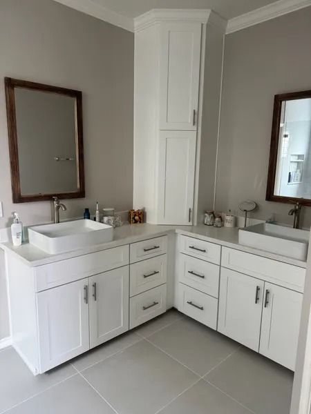 White bathroom with two sinks, two mirrors, white cabinets, corner storage, and light gray walls.