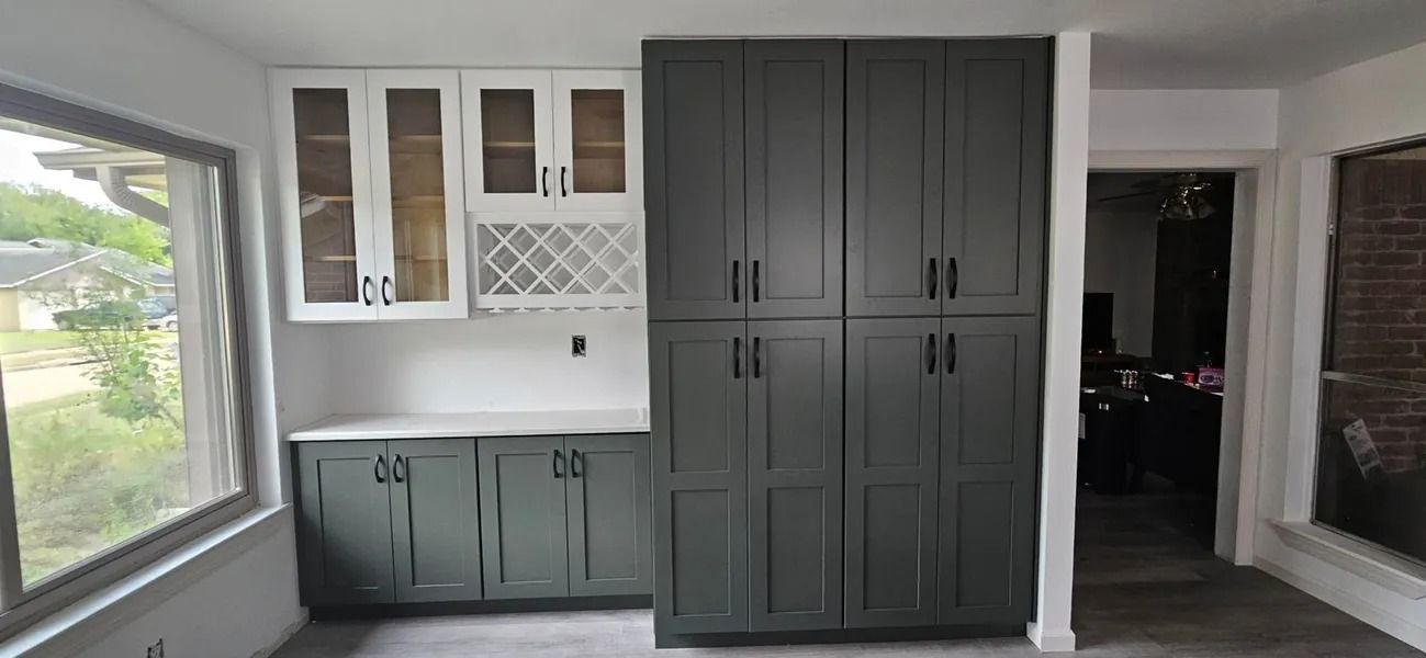 A kitchen with white cabinets, dark green lower cabinets, and dark gray pantry cabinets, next to a window.
