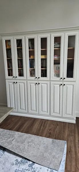 White cabinets with glass doors, and dark handles. Brown wooden floor. Grey rug on floor.