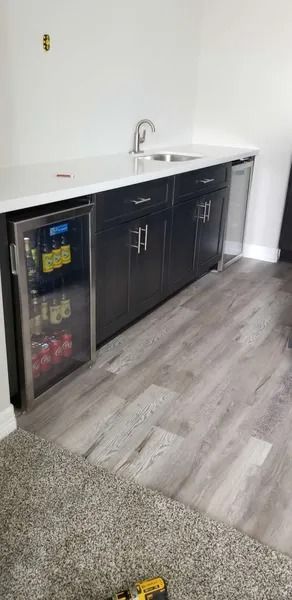 A home wet bar with a mini fridge, sink, and cabinets. Gray flooring and white countertop.