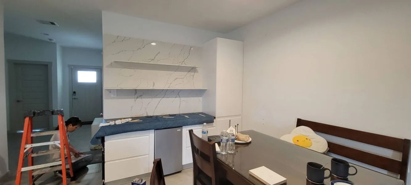 Kitchen with marble backsplash, white cabinets, and a dining table. A person works on a ladder.