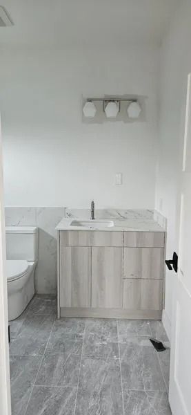 Bathroom with gray tiled floor, white walls, light wood vanity, and a toilet.