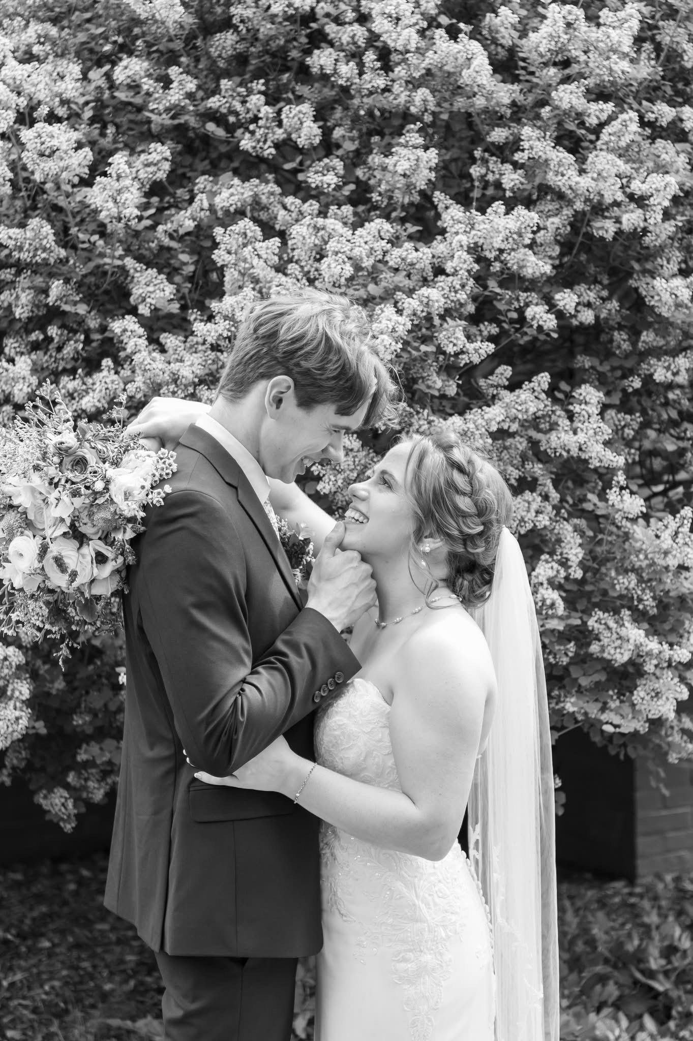 Newlywed couple embracing, smiling at each other. The bride wears a strapless gown and veil, the groom a suit, set against a backdrop of flowering foliage.
