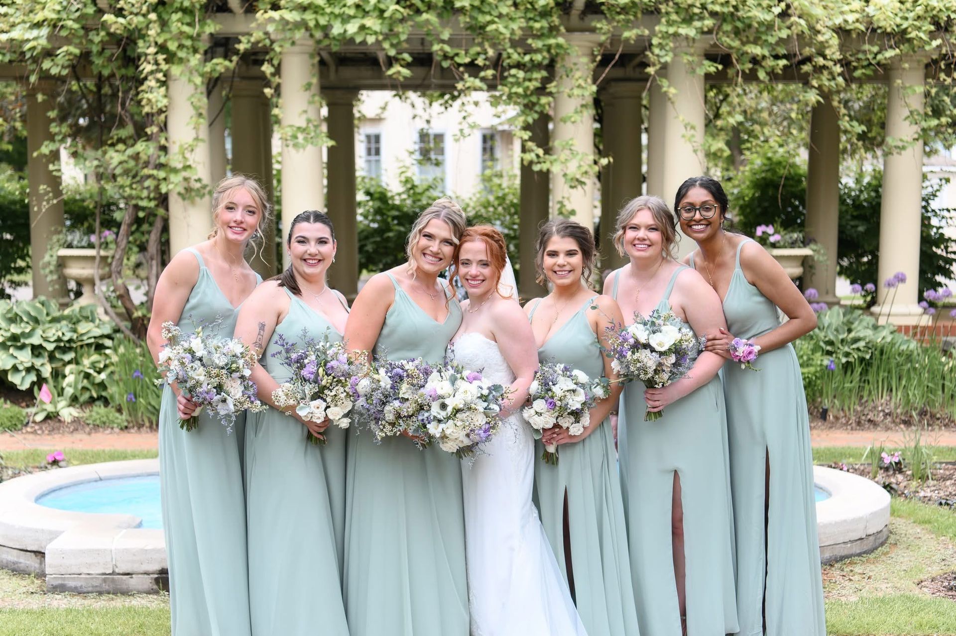 A bride with red hair and six bridesmaids in sage green dresses pose outdoors. They hold bouquets in a garden setting.