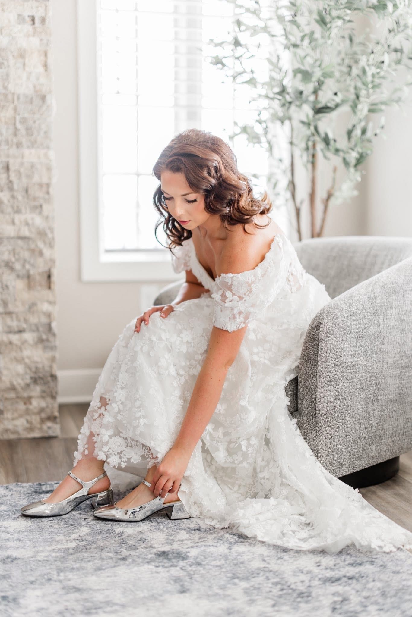 A bride in a white floral gown puts on her silver shoes indoors, seated on a gray couch.