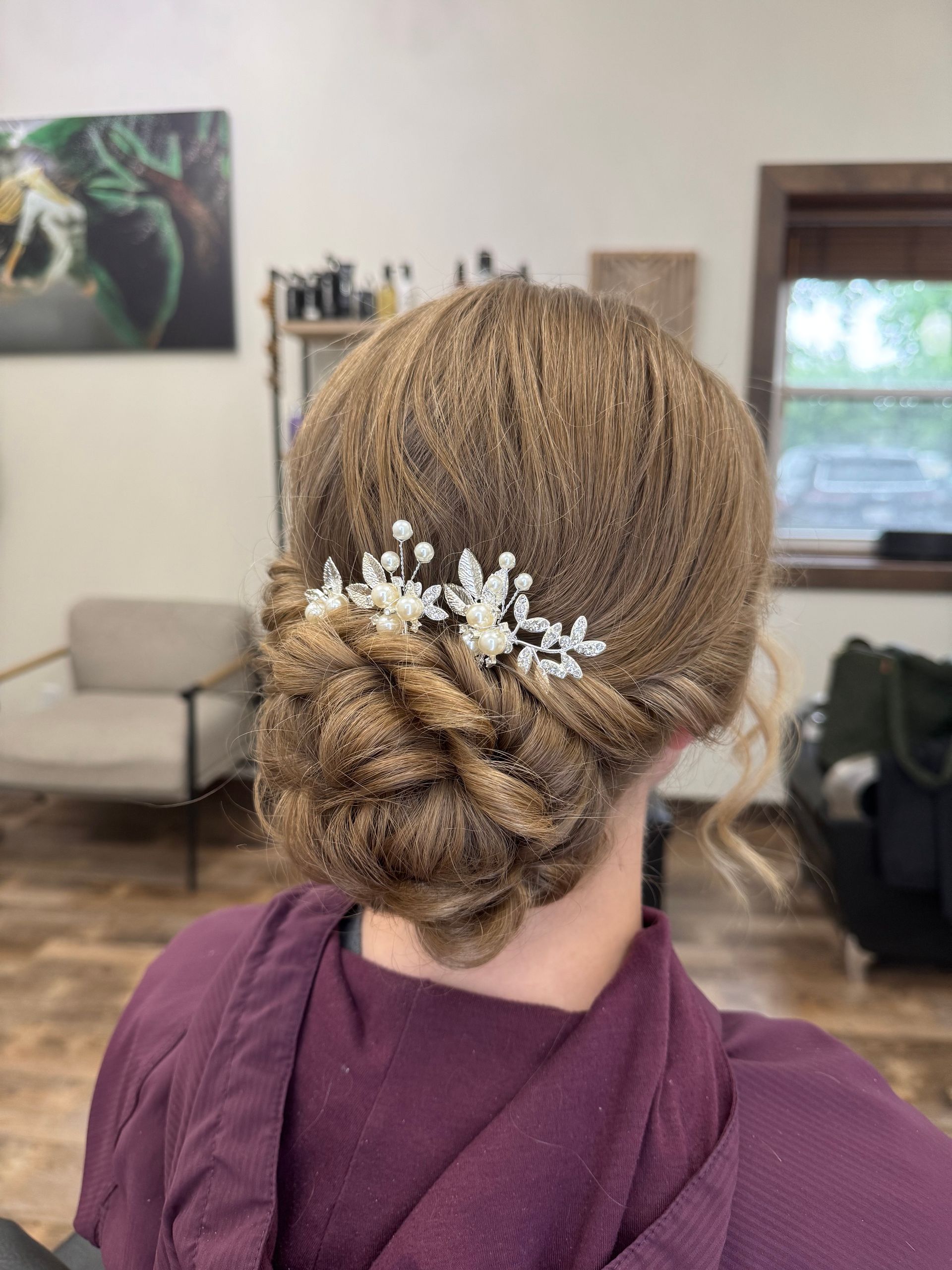Woman with light brown hair styled in a low bun with a decorative hairpiece. She wears a purple robe indoors.