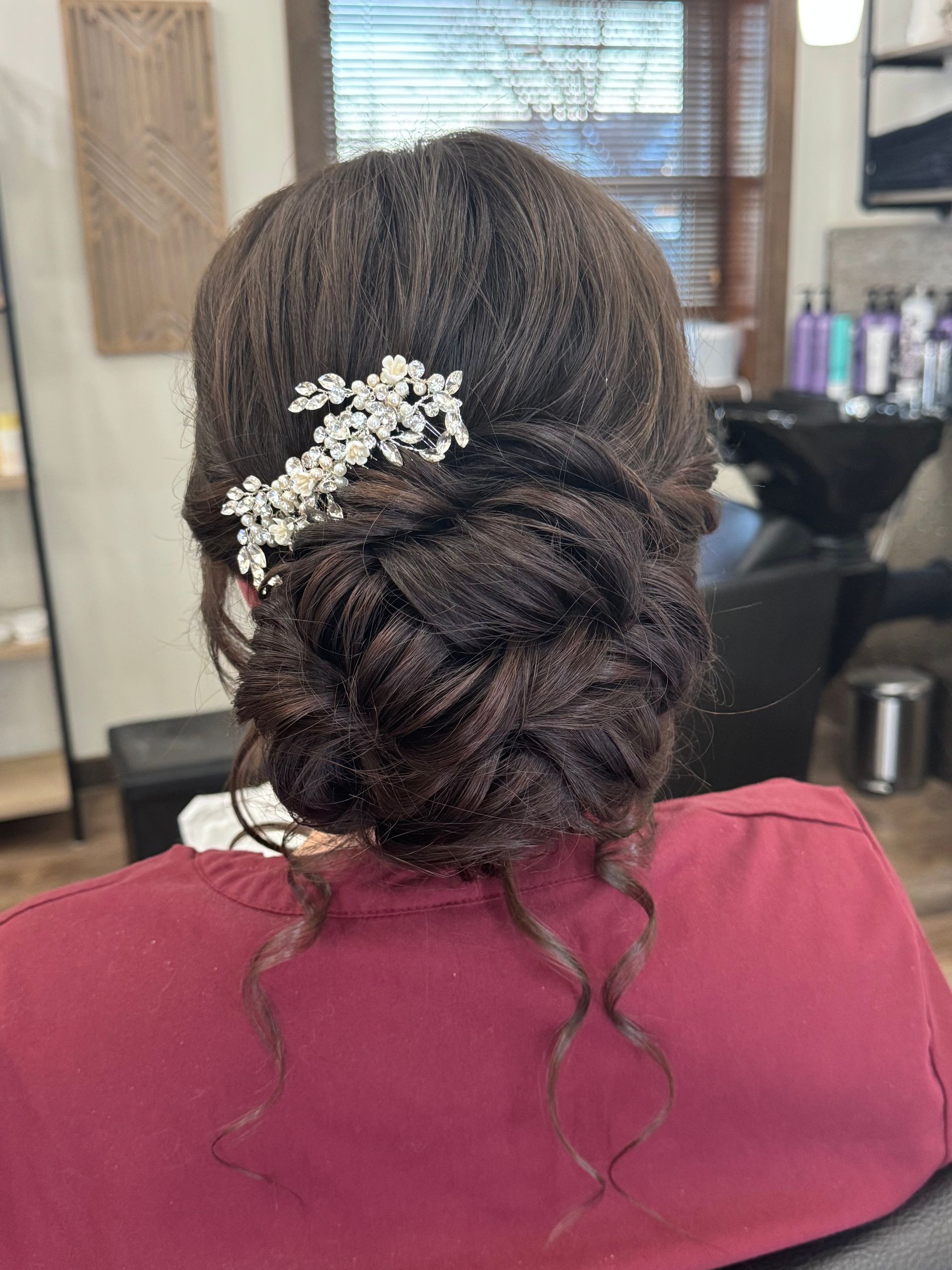 Woman with dark hair styled in an updo, adorned with a jeweled hairpiece. The hairstyle sits against a salon backdrop.