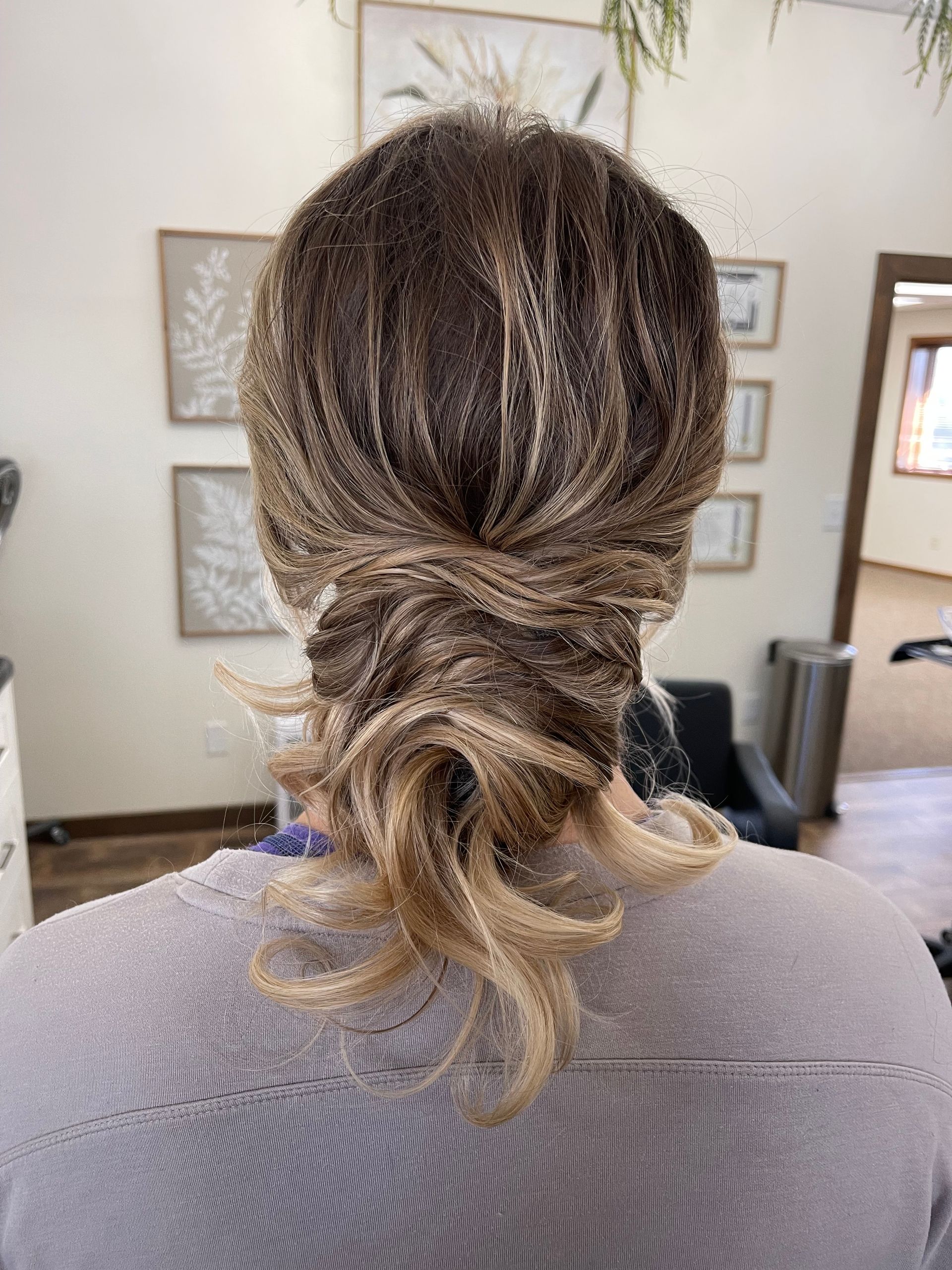 Woman with dark brown and blonde hair styled in a low bun, viewed from the back. She wears a gray sweater, indoors near framed art.