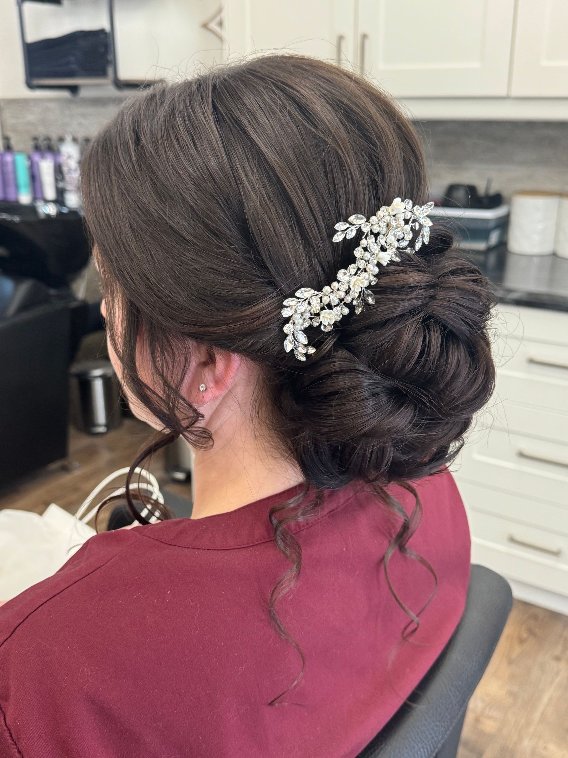 Woman with dark brown hair styled in a low bun, adorned with a silver hairpiece. She's wearing a maroon shirt, indoors in a salon.